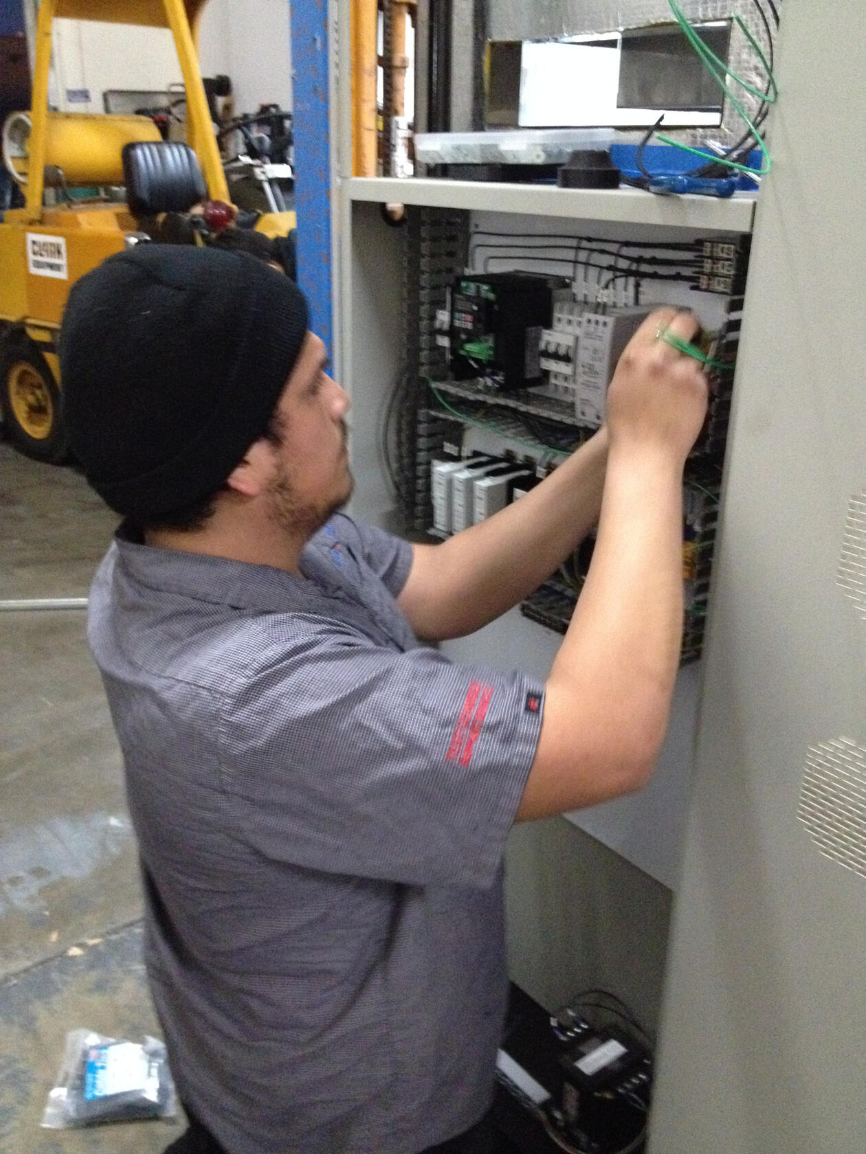 Technician Performing Electrical Maintenance A skilled technician working on the internal wiring of an electrical panel, carefully adjusting components. The technician wears a beanie and a checkered uniform, with tools and equipment visible in the background.